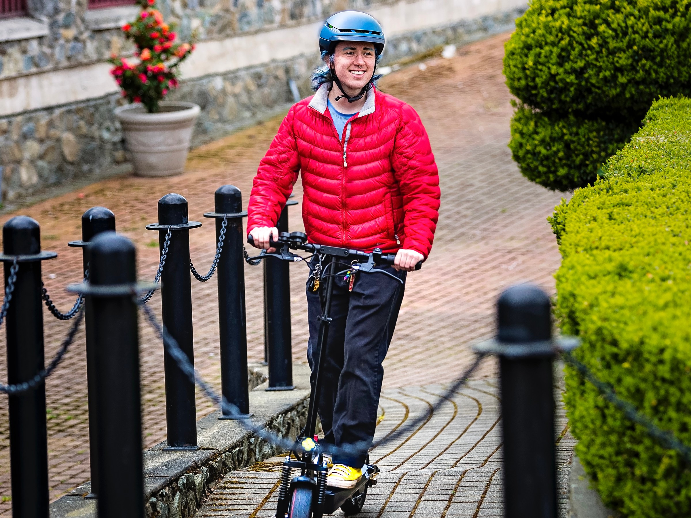 Person in red jacket and helmet riding an electric scooter on a cobblestone path.