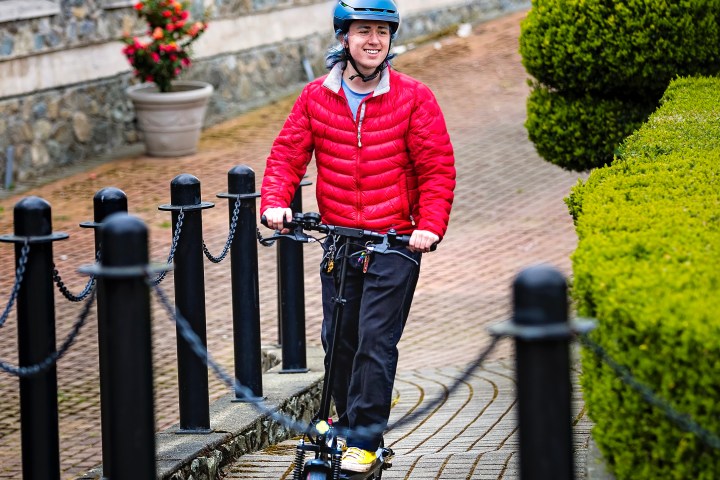 Person in red jacket and helmet riding an electric scooter on a cobblestone path.