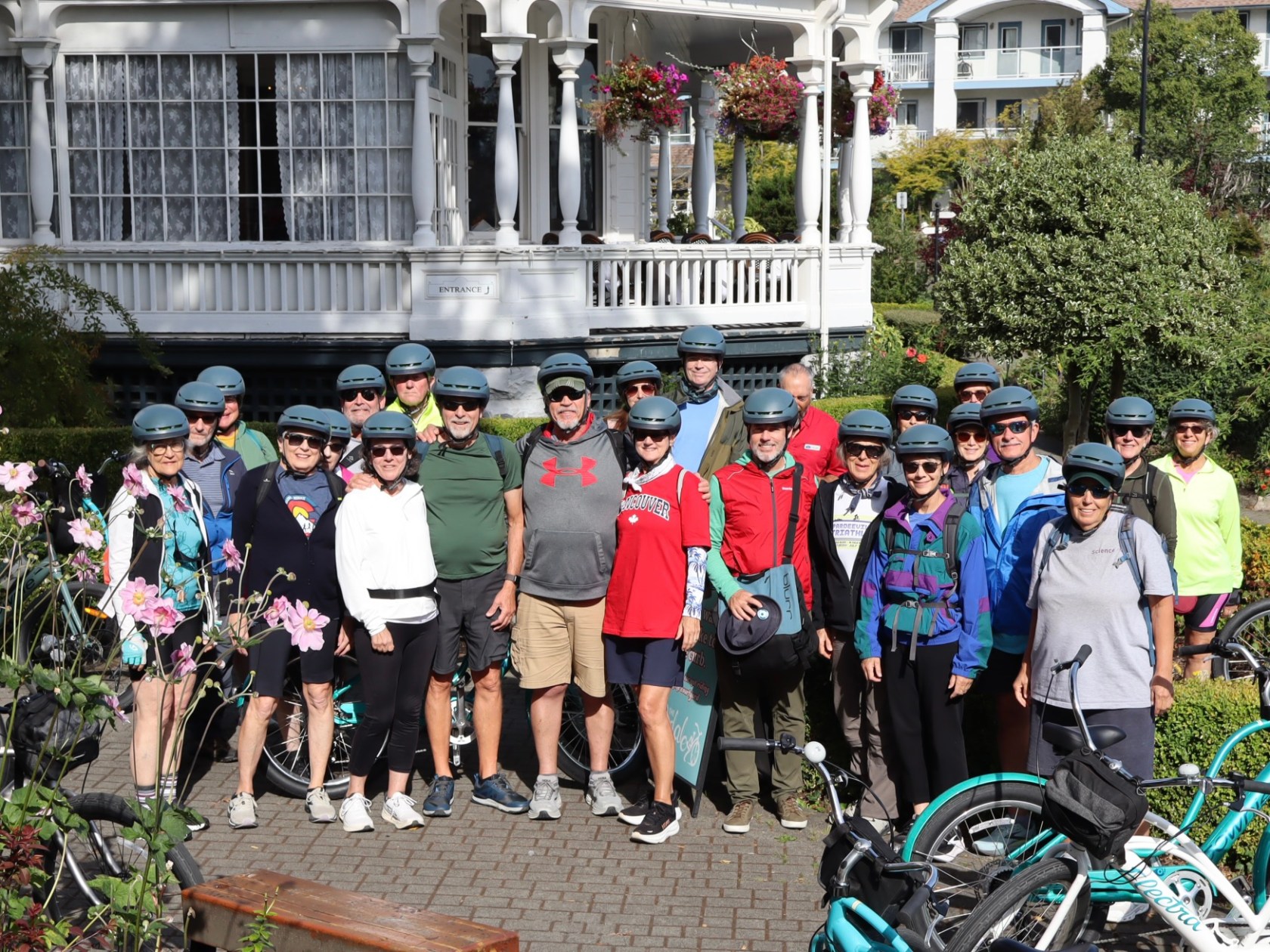 Group of people with helmets and bikes posing in front of a white building with flowers.