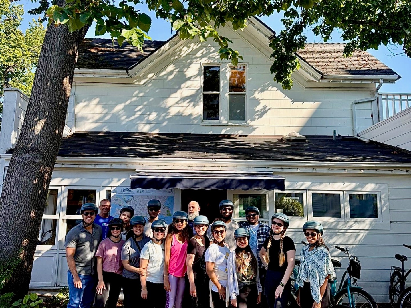 Group of cyclists with helmets posing in front of a white house under a large tree.